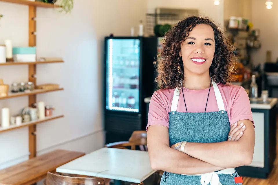 A café owner in a pink T-shirt and apron smiling behind her counter