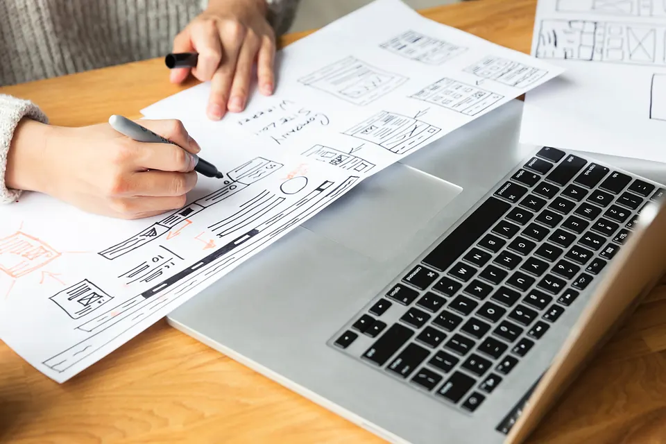 Hands sketching a website layout on paper next to a laptop